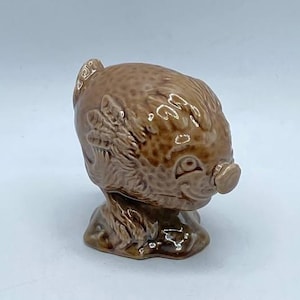 May include: A small, glossy, light brown ceramic figurine of a stylized pig. The figure features a rounded body with textured, leaf-like patterns carved into the surface, a prominent snout, and a smooth, glazed finish that reflects light.
