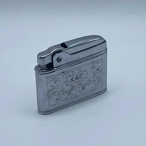 May include: A silver-colored lighter with an engraved floral design and the initials "A.M.G." The lighter has a rectangular body with a hinged top. The lighter is shown against a white background.