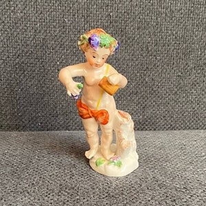 May include: A porcelain figurine of a young boy wearing a wreath of flowers and holding a small basket. The boy is standing on a tree stump and is painted in shades of pink, orange, and green.