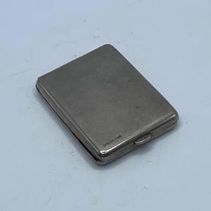 May include: A rectangular, silver-colored cigarette case with a hinged lid. The case is made of metal and has a smooth, reflective surface. The case is closed and appears to be empty. The case is a vintage item.