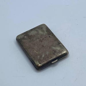 May include: A rectangular, antique silver cigarette case with a slightly tarnished finish. The case has a hinged lid and a small, curved clasp. The overall design is simple and classic, suggesting a vintage aesthetic.