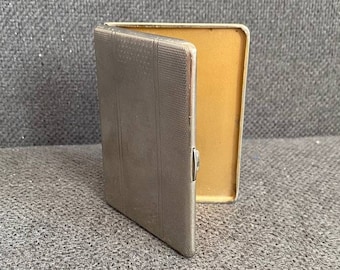 May include: A silver cigarette case with a textured design and a hinged lid. The case is open, revealing a gold interior.
