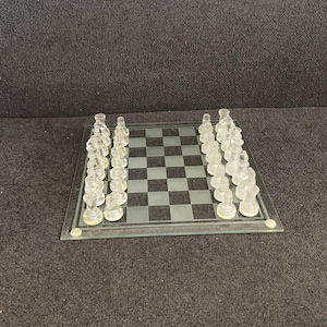 Könnte beinhalten: Ein Schachspiel aus klarem Glas mit einem karierten Schachbrett. Die Schachfiguren sind transparent und auf dem Brett angeordnet. Das Brett ist quadratisch und liegt auf einer dunklen Oberfläche.