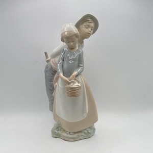 Può includere: Statuina in porcellana raffigurante una ragazza con un cestino, con un ragazzo che guarda sopra la sua spalla. La ragazza indossa un vestito grigio, un grembiule bianco e un cappello. Il ragazzo indossa un cappello e una camicia chiara. Colori pastello.