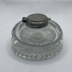 May include: A clear glass inkwell with a round, domed shape and a decorative, ridged base. The inkwell has a hinged, metal lid. The glass is transparent, allowing a view of the interior. The inkwell is a vintage writing accessory.