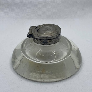 May include: A clear glass inkwell with a round, shallow base and a hinged metal lid. The lid is circular with a decorative design. The inkwell is likely vintage and sits on a white surface.