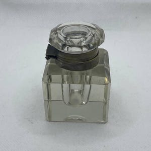 Puede incluir: Un tintero de vidrio transparente con una tapa decorativa facetada. El tintero es de forma cuadrada con una banda de metal alrededor de la parte superior. La tapa es un diseño transparente y multifacético. Es un artículo vintage.