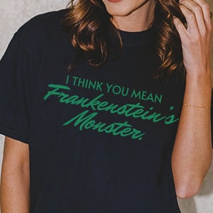 May include: Black t-shirt with the text "I THINK YOU MEAN Frankenstein's Monster" in green script. The shirt is a dark color and has a classic crew neck design. The text is centered on the front of the shirt.