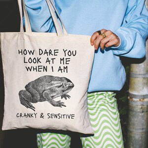 May include: A white canvas tote bag with black text that reads "How dare you look at me when I am cranky & sensitive". A black and white illustration of a frog is centered on the bag.