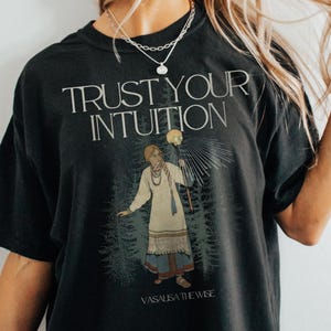 Puede incluir: Camiseta negra con un estampado gráfico de una mujer sosteniendo un bastón y el texto "TRUST YOUR INTUITION" y "VASALISA THE WISE".
