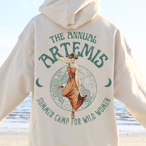 May include: A cream-colored hoodie with a graphic print of a woman holding a bow and arrow. The graphic includes the text "The Annual Artemis Summer Camp for Wild Women".