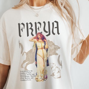 Puede incluir: Una camiseta blanca con un estampado gráfico de una mujer con un vestido blanco de pie junto a un lobo. El texto "FREYA" está impreso en negro sobre el gráfico. El texto "To be blessed by the Goddess of Magic, Sorceres, Secres, and Down-Bringer, The Gift Giver, Warrior, and Goddess of Love." está impreso debajo del gráfico.