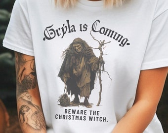 Puede incluir: Camiseta blanca con una ilustración en negro y marrón de una mujer con un bastón y una cesta. El texto "Gryla is Coming. Beware the Christmas Witch." está impreso en la camiseta.