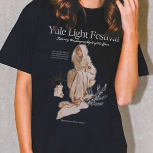 May include: A dark blue t-shirt with a graphic print of a woman in a white robe holding a candle. The text on the shirt reads "Yule Light Festival Starring the Longest Night of the Year" and "Honoring the Winter Solstice".