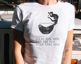 May include: White crop top with a black graphic of a mermaid and seal. The text on the shirt reads "STEAL HER SKIN, AND THE SEA WILL TAKE YOU." The shirt is short-sleeved and has a crew neck.