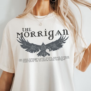 May include: Cream-colored t-shirt featuring the text "THE MORRIGAN" above a stylized raven graphic. Below the raven, text lists titles associated with the Morrigan. The shirt is a relaxed fit.