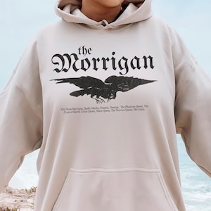 May include: A beige hooded sweatshirt with a black graphic print of a raven and the text "the Morrigan". The text below the raven reads "The Thew Morrigss, Bad Macha, Nomain, Dunsus, The Phantom Queen, The Crow of Battle, Great Queen, Mars-Quen, The Warrior Queen, Morrigas."