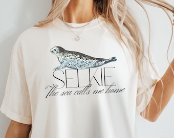 May include: Cream-colored t-shirt featuring a graphic of a seal with the word "SELKIE" above it. Below the seal, the text reads "The sea calls me home" in a cursive font. The seal is depicted in shades of gray and blue.