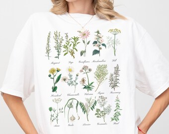 Puede incluir: Camiseta blanca con un estampado botánico que presenta ilustraciones de varias hierbas y flores. Los nombres de cada planta están impresos debajo de la imagen, incluyendo: "Mejorana", "Salvia", "Aciano", "Malvavisco", "Eneldo", "Consuelda", "Manzanilla", "Valeriana", "Hisopo", "Romero", "Angélica", "Ajo", "Apio", "Cilantro", "Albahaca"