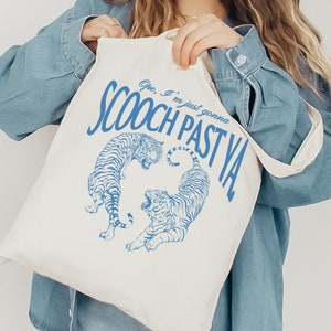 Peut inclure: Sac fourre-tout blanc cassé avec le texte bleu "SCOOCH PAST YA" et deux illustrations de tigres. Le sac a un design simple avec une forme rectangulaire et une longue sangle pour le porter à l'épaule.