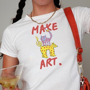May include: White t-shirt with the words "MAKE ART" in red, featuring a whimsical illustration of a cat and a spotted animal. The shirt is paired with a red shoulder bag and a gold necklace. A martini glass with olives is held in the hand.