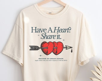May include: A beige t-shirt with a graphic of two red hearts pierced by an arrow. The text "Have A Heart? Share it." is above the graphic. The text "Become an Organ Donor. Apply online with your state's registry or with the MVD." is below the graphic.