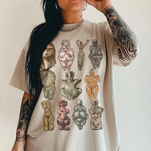 Può includere: T-shirt beige oversize con un'illustrazione colorata di varie antiche figure femminili. L'opera d'arte comprende statue nei toni del beige, rosa e marrone. La maglietta ha maniche corte e una vestibilità ampia.