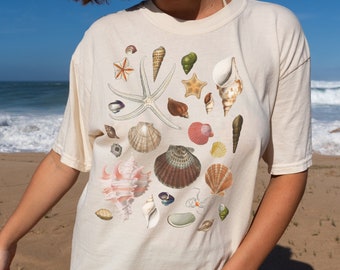 Puede incluir: Una camiseta blanca con un estampado de conchas marinas de colores. El estampado presenta una variedad de conchas, estrellas de mar y otras imágenes con temática oceánica.