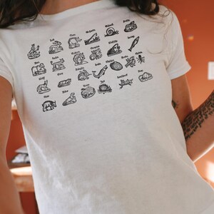 May include: White t-shirt featuring a grid of black illustrations of various animals, each labeled with a corresponding name. The shirt is made of a soft, light-colored fabric.