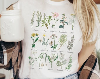Puede incluir: Una camiseta blanca con un estampado botánico de quince hierbas y flores, incluyendo artemisa, salvia, equinácea, malvavisco, eneldo, cola de caballo, manzanilla, valeriana, tomillo, romero, anís, ajo, regaliz, cilantro y albahaca.
