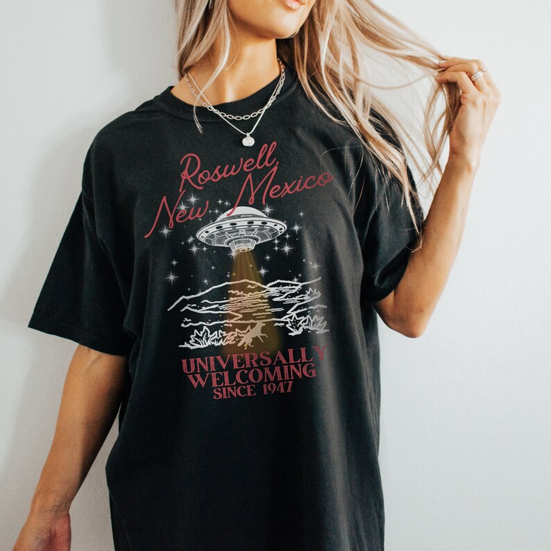 Puede incluir: Camiseta negra con un gr&aacute;fico de un OVNI sobre un paisaje des&eacute;rtico. El texto "Roswell New Mexico Universally Welcoming Since 1947" est&aacute; impreso en la camiseta.