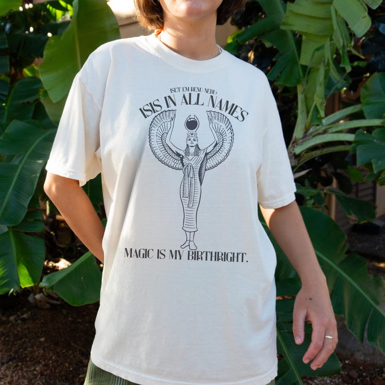 May include: Cream-colored t-shirt featuring a black graphic of the Egyptian goddess Isis with outstretched wings. The text "ISIS IN ALL NAMES" and "MAGIC IS MY BIRTHRIGHT" are printed above and below the graphic, respectively.
