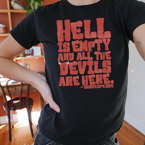 Puede incluir: Camiseta negra con la cita "Hell is Empty and All the Devils Are Here" en letras rojas, atribuida a Shakespeare. La camiseta es de cuello redondo.