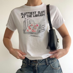 May include: White t-shirt with the text "Sluttiest Slut at the Library" above a graphic of books and lipstick kisses. The shirt is paired with blue jeans and a black shoulder bag.