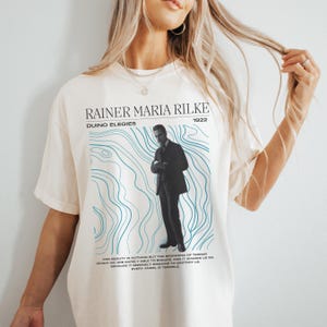 May include: A cream-coloured t-shirt with a black and white image of a man, and the text "RAINER MARIA RILKE 1922 DUINO ELEGIES". Below the image are blue wavy lines and a quote. A casual, unisex tee.