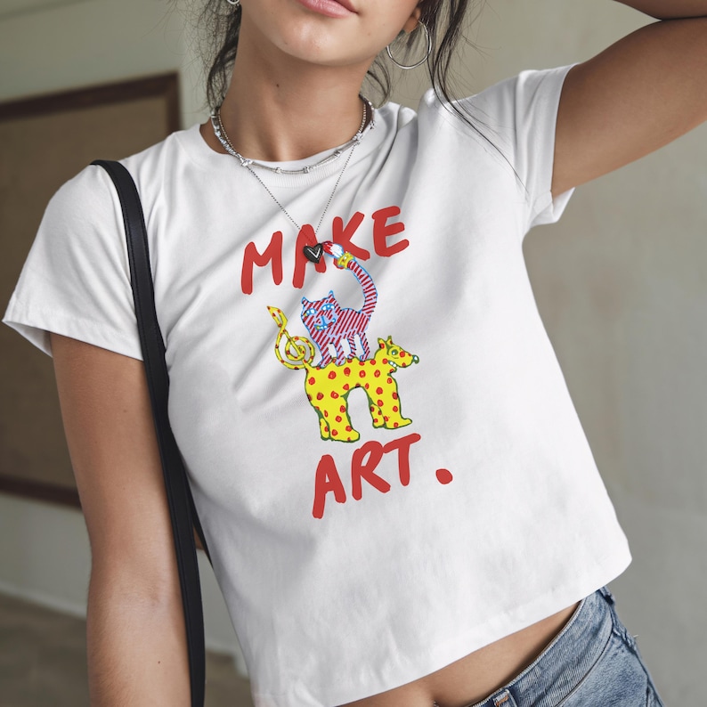 May include: White t-shirt with the words "MAKE ART" in red. The shirt features a colorful graphic of a cat and a dog. The cat is pink and blue, and the dog is yellow with red spots.