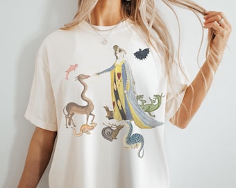 Op de afbeelding: Een wit T-shirt met een kleurrijke grafische print van een vrouw in een lange jurk omringd door verschillende mythische wezens, waaronder een draak, een eenhoorn en een slak.