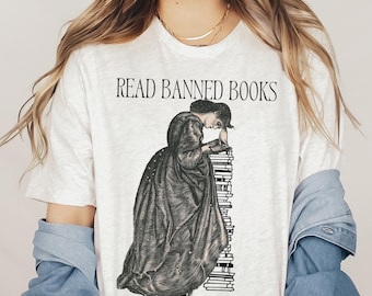 May include: A white t-shirt with the text "READ BANNED BOOKS" and an illustration of a woman reading a stack of books. The text "GAIN FORBIDDEN KNOWLEDGE" is printed below the illustration.