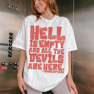 Puede incluir: Camiseta blanca con la cita "HELL IS EMPTY AND ALL THE DEVILS ARE HERE." en rojo, atribuida a Shakespeare. La camiseta tiene un corte holgado y mangas cortas. El texto está en negrita, con un estilo retro.