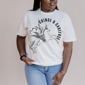 May include: Cream-colored t-shirt featuring a black graphic of a winged horse ridden by a figure with a sword. The words "CRINGE & CAREFREE" are arched around the image. The shirt is made of a soft material.
