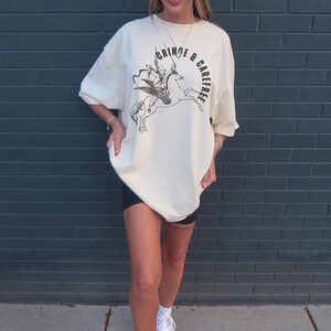 May include: Cream-colored oversized t-shirt with a black graphic of a winged figure riding a horse, and the text "CRINGE & CAREFREE". The shirt is paired with black shorts and white sneakers.