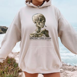 Puede incluir: Sudadera con capucha beige con un estampado de "Calavera con un cigarrillo encendido" de Vincent van Gogh. La obra de arte está centrada en la parte delantera, con el nombre del artista y el título debajo. La sudadera tiene un bolsillo de canguro y una capucha con cordón.