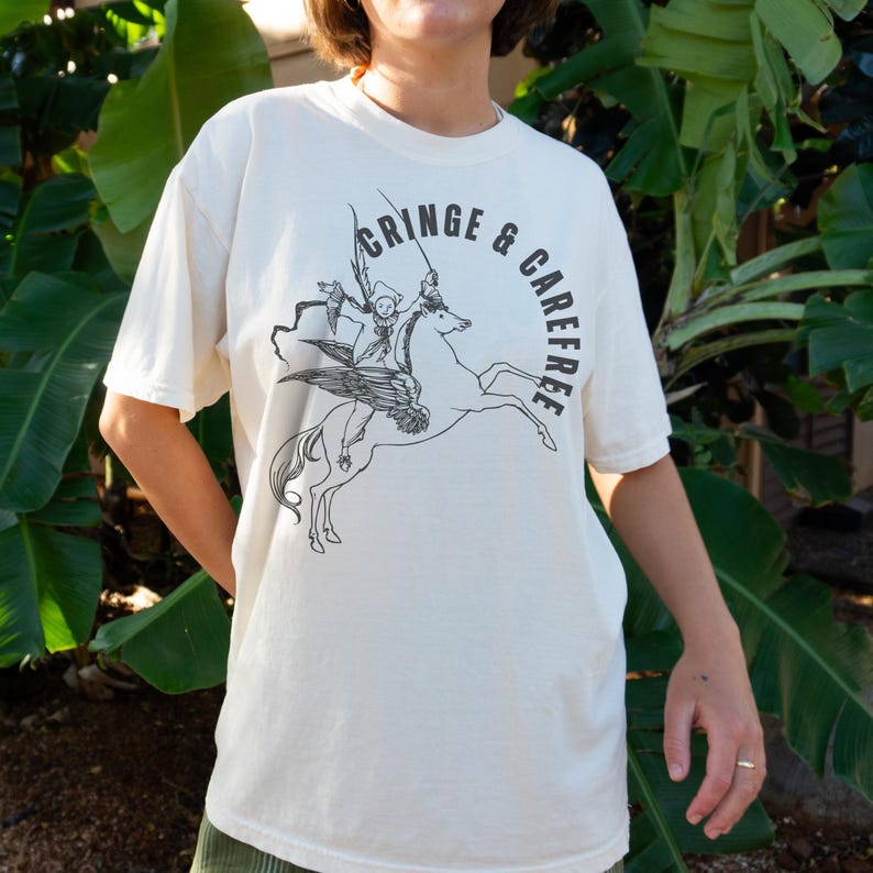 May include: Cream-colored t-shirt with a black graphic of a winged horse ridden by a skeleton figure. The words "CRINGE & CAREFREE" are printed in a circle around the image. The shirt has short sleeves and a crew neck.