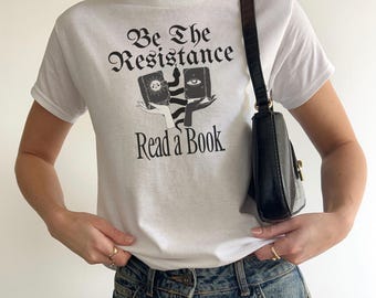 May include: White t-shirt with the text "Be The Resistance Read a Book" in black script. The graphic features two hands holding books with mystical symbols. A black purse with a gold clasp hangs from the shoulder.
