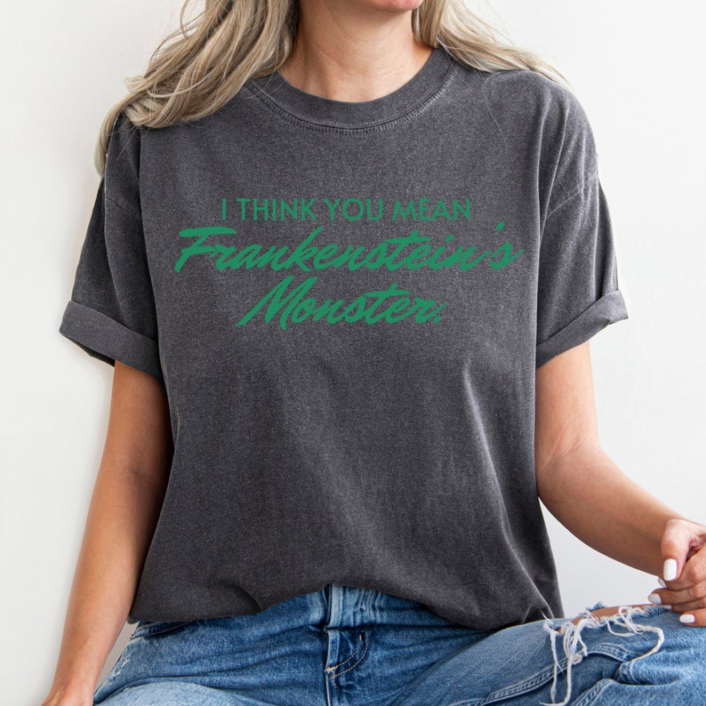 May include: Dark gray t-shirt with rolled-up sleeves. The shirt features the text "I THINK YOU MEAN Frankenstein's Monster" in green script. The shirt is a casual style, suitable for everyday wear.