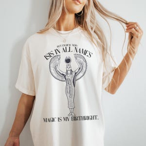 May include: Cream-colored t-shirt featuring a black graphic of an Egyptian goddess with outstretched wings, the text "ISIS IN ALL NAMES" above, and "MAGIC IS MY BIRTHRIGHT" below. The shirt has short sleeves and a relaxed fit.