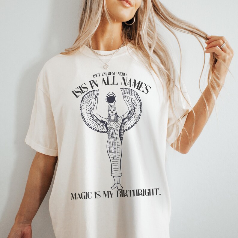 May include: Cream-colored t-shirt featuring a black graphic of an Egyptian goddess with outstretched wings, the text "ISIS IN ALL NAMES" above, and "MAGIC IS MY BIRTHRIGHT" below. The shirt has short sleeves and a relaxed fit.