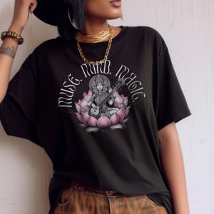 May include: Black t-shirt with a detailed illustration of a deity playing a stringed instrument, seated on a pink lotus flower. The text "Muse. Mind. Magic" arches above the illustration in a stylized font.