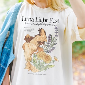 以下が含まれることがあります： 花に囲まれた女性のグラフィックプリントが施された白いTシャツ。グラフィックの上に「Litha Light Fest」というテキストが印刷されています。グラフィックの下に「Starring the Longest Day of the Year」というテキストが印刷されています。グラフィックの下に「Midsummer has come so watch the rise and set of the Sun now that it is at it's full power.」というテキストが印刷されています。グラフィックの下に「Celebrating on the Summer Solstice.」というテキストが印刷されています。