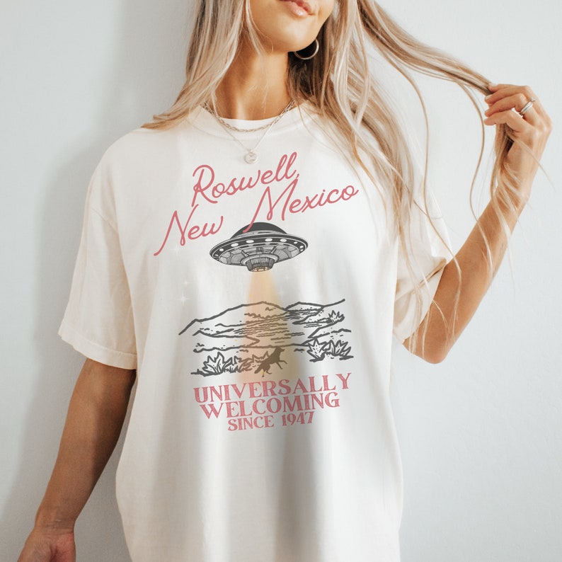 Puede incluir: Una camiseta blanca con un dise&ntilde;o gr&aacute;fico de un OVNI volando sobre un paisaje des&eacute;rtico. El texto "Roswell New Mexico" est&aacute; escrito en cursiva rosa sobre el OVNI. El texto "Universally Welcoming Since 1947" est&aacute; escrito en rosa debajo del paisaje.